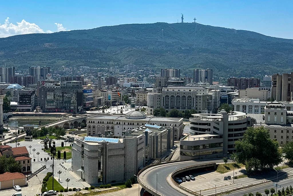 Panoramic view of Skopje city center with Mount Vodno and the Millennium Cross in the background, featuring the Holocaust Memorial Center and Stone Bridge